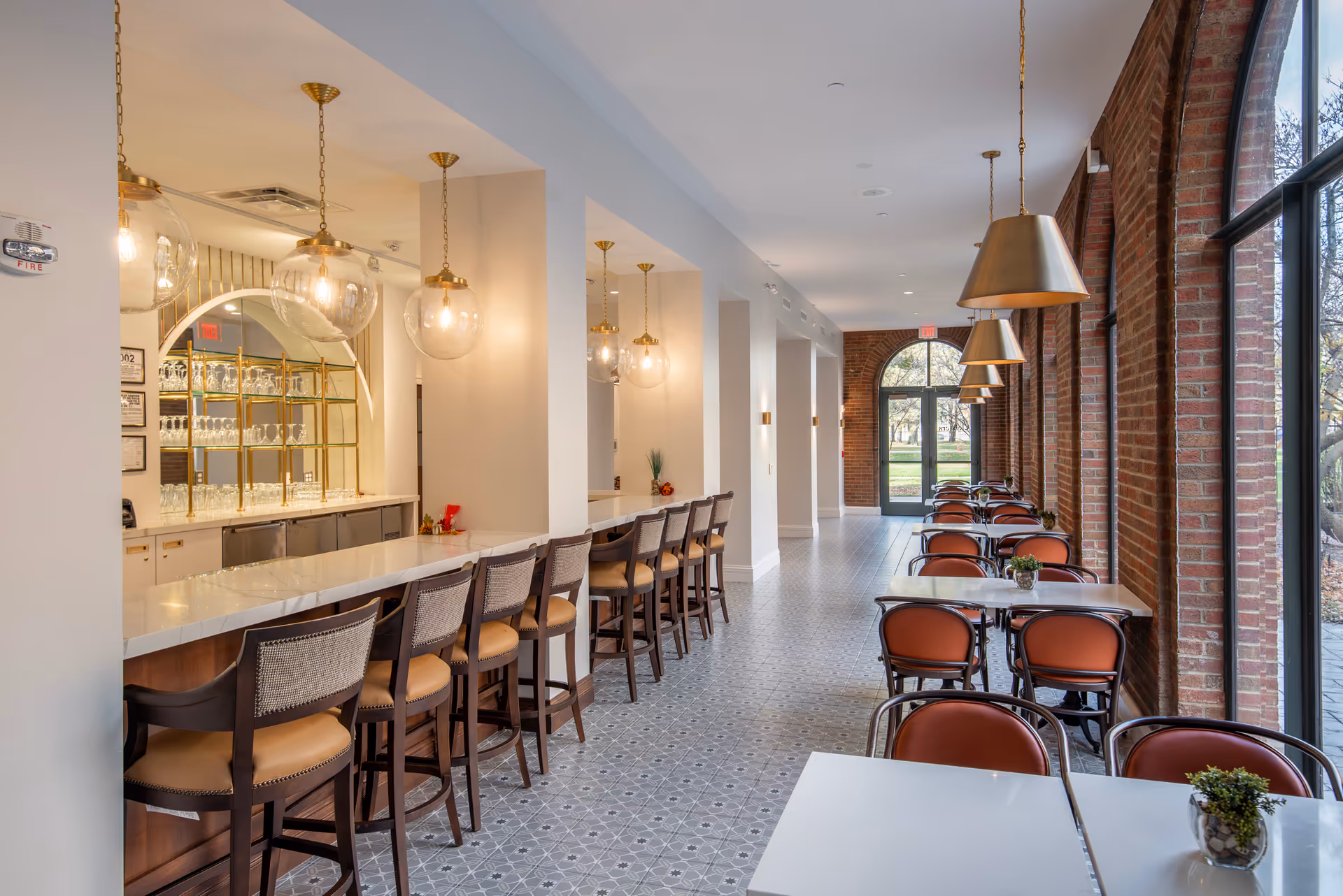 Bright dining area with a long marble bar and barstools on the left and rows of tables and chairs along large arched windows on the right.
