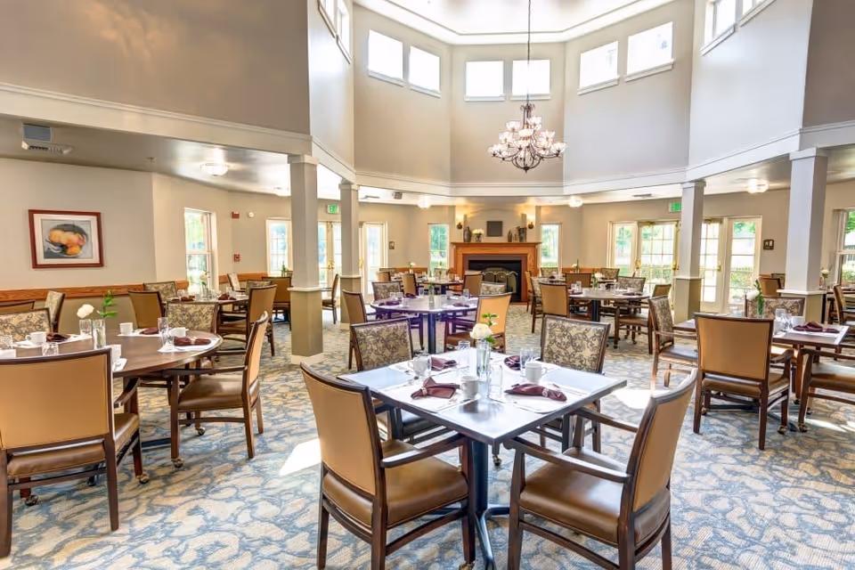 A bright and spacious dining room with multiple tables set with cups, glasses, napkins, and small flower vases. The room features large windows allowing natural light, a chandelier hanging from a high ceiling, and a fireplace at the far end. Chairs with cushioned seats and backs surround the tables, and the carpet has a patterned design.