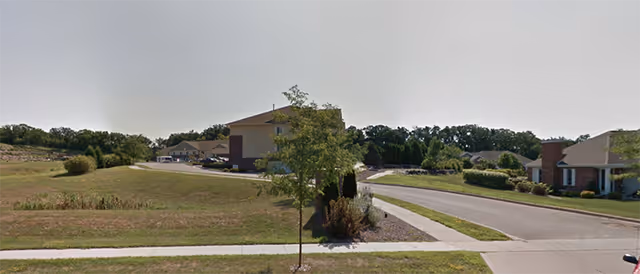 A suburban street view showing a few residential buildings with well-maintained lawns and trees under a clear sky.