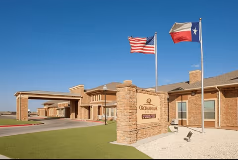 Front entrance of Orchard Park of Permian Basin with a stone sign, flagpoles, and a covered drive.
