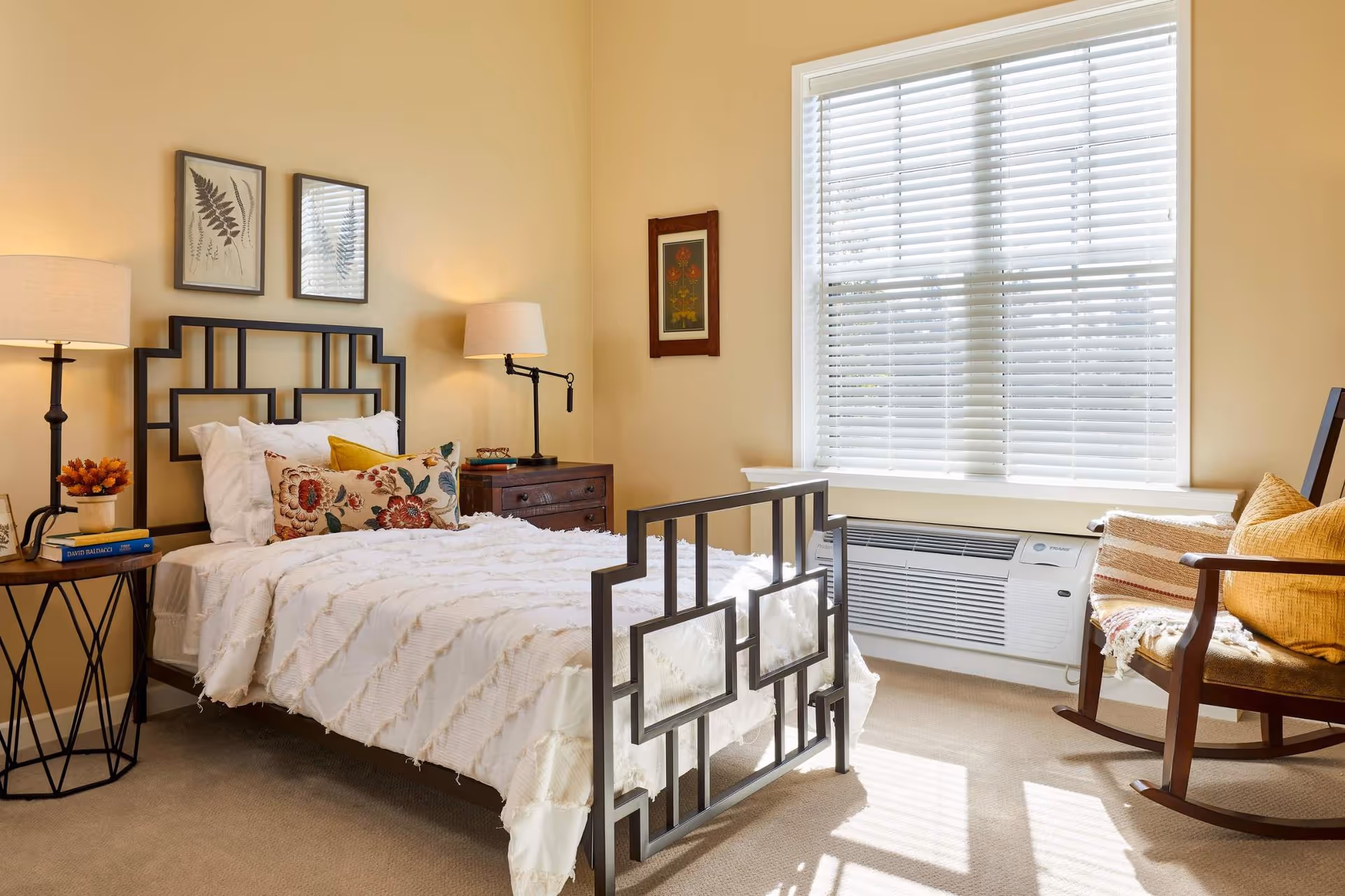 Sunlit bedroom with a decorative metal bed, bedside tables and lamps, a window with blinds and a rocking chair.