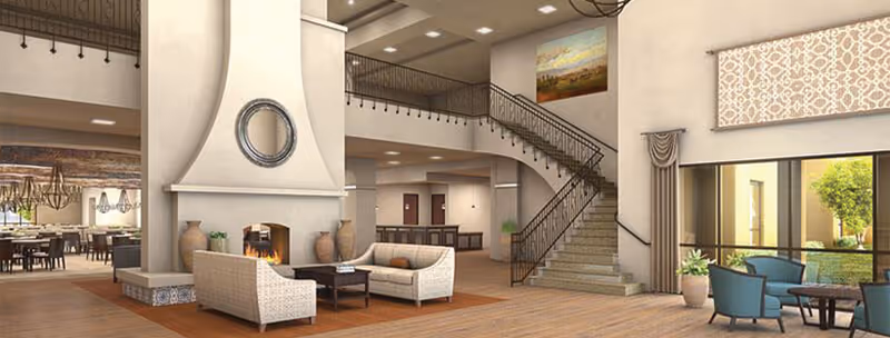 Bright senior living lobby with seating around a fireplace, a curved staircase, and a dining area visible in the background.