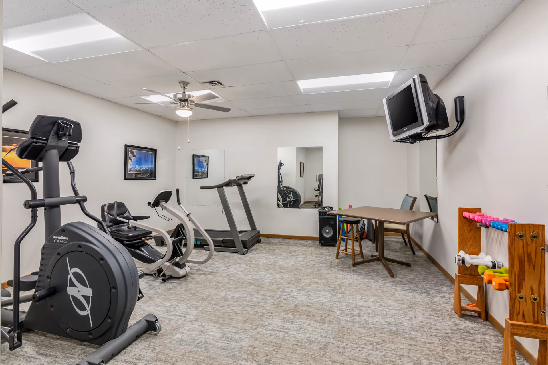 A senior living facility exercise room with various fitness equipment including a recumbent bike, treadmill, elliptical machine, and a rack of colorful hand weights. The room has a ceiling fan, fluorescent lighting, a wall-mounted TV, mirrors on the walls, a small table with a stool, and framed pictures on the walls.