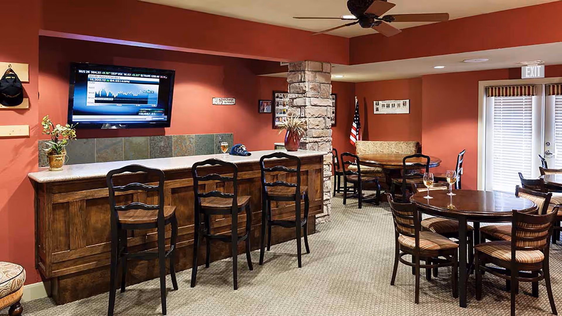 Interior view of a senior living facility lounge area with a wooden bar counter and three high chairs. A flat-screen TV is mounted on the red wall above the bar. There are round tables with chairs and glasses of white wine on them. A stone pillar and an American flag are visible in the background, along with a cushioned bench seat. The room has carpeted flooring and a ceiling fan.