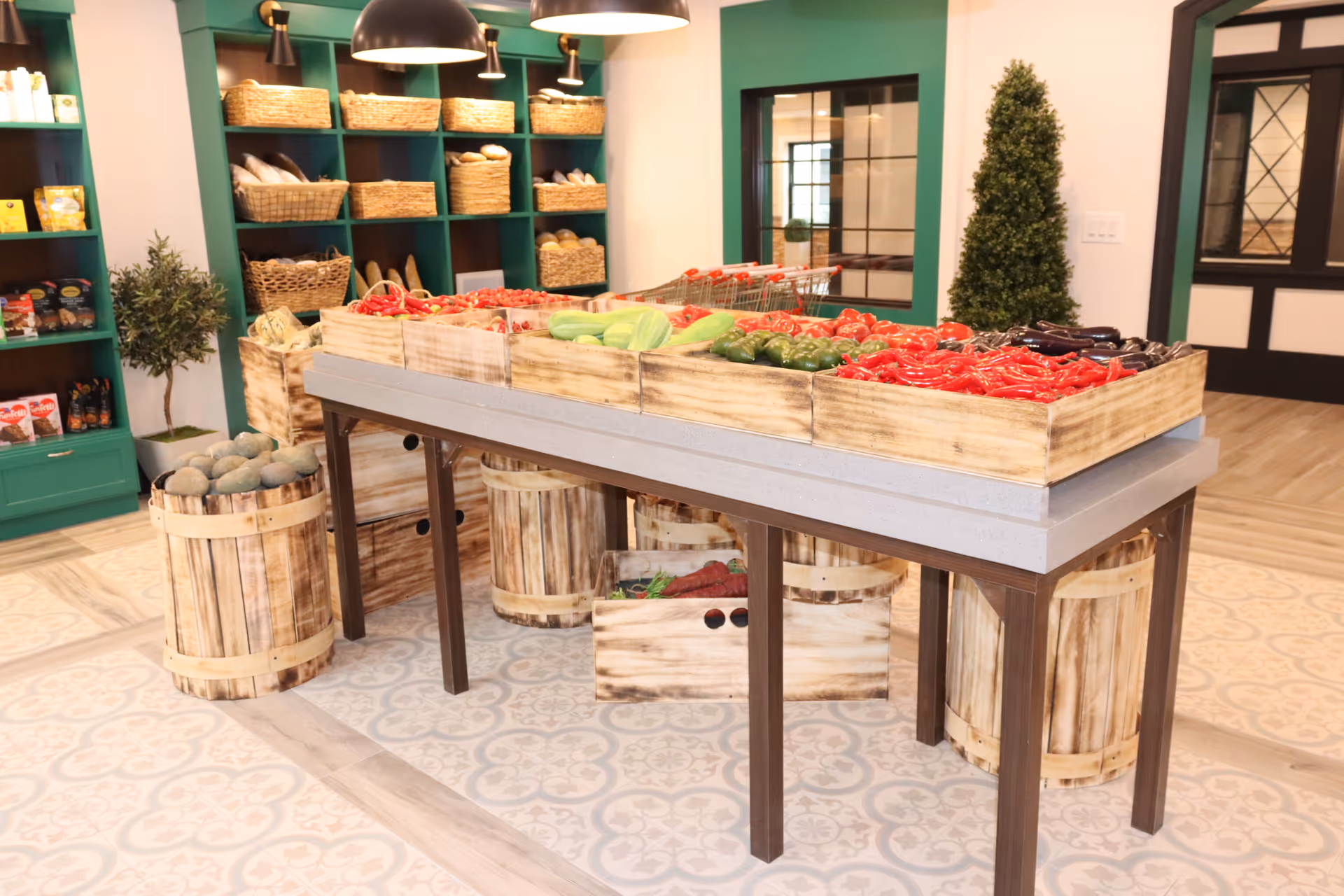 Interior of a facility room with a wooden table displaying fresh vegetables such as red peppers, green peppers, and eggplants in wooden crates. Surrounding the table are wooden barrels filled with produce. Shelves in the background hold baskets of bread and other food items. The room has patterned tile flooring, green shelving units, and decorative plants.
