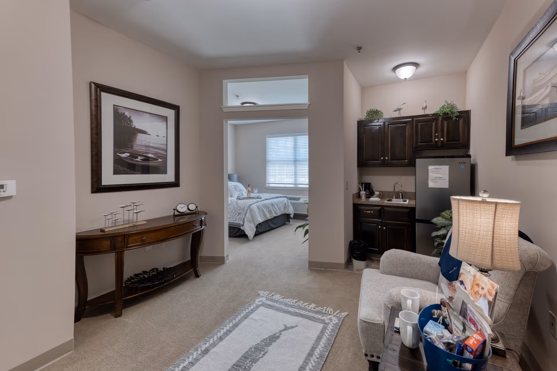 Interior view of a senior living facility apartment showing a small living area with a beige armchair, a side table with a lamp and various items, a kitchenette with dark wood cabinets, a mini refrigerator, and a sink. A hallway leads to a bedroom with a bed, window with blinds, and a bottle of wine on a tray. The walls are decorated with framed pictures, and a wooden console table with decorative items is against one wall.
