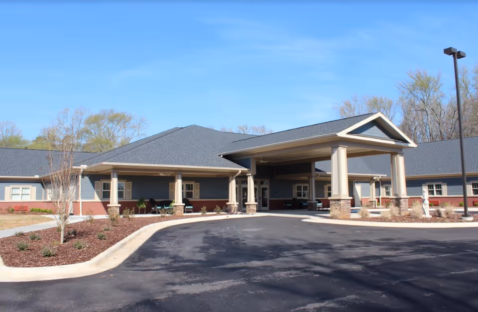 Exterior view of Heritage Assisted Living & Memory Care facility showing a single-story building with a covered entrance supported by large columns, surrounded by landscaped areas and a paved driveway under a clear blue sky.
