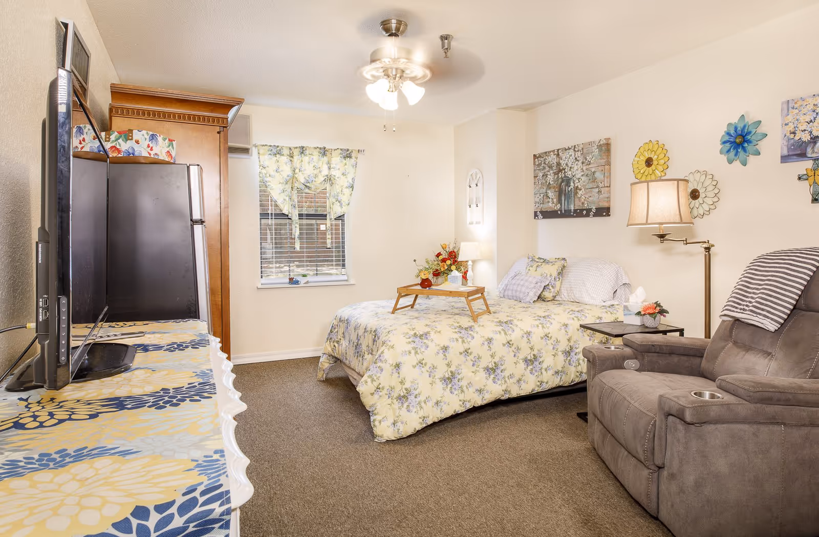 Sunny furnished senior bedroom with floral bedding, a recliner, TV on a dresser, and wall decorations.