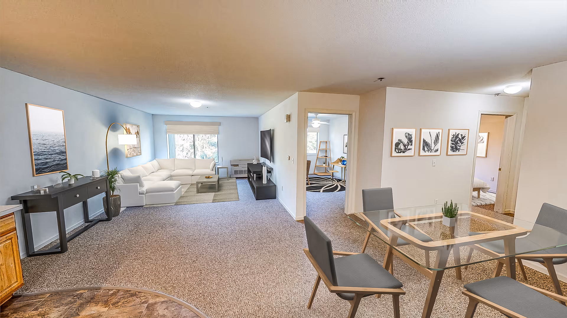 A spacious and well-lit living area in an apartment or senior living facility. The room features a white sectional sofa with a coffee table in front, a flat-screen TV mounted on a stand, and a console table with decorative items along one wall. There is a glass dining table with four chairs in the foreground. The walls are decorated with framed artwork, and there are windows letting in natural light. The carpeted floor and neutral color scheme create a cozy and inviting atmosphere.