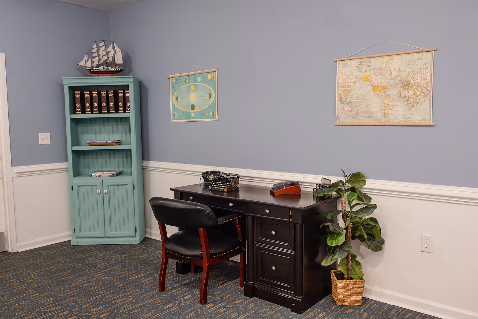 A small office or study area with a black wooden desk and a black leather chair. On the desk are a vintage typewriter and a wooden organizer. To the left is a light blue bookshelf with several books and a model ship on top. The walls are painted light blue with white wainscoting, and two vintage-style maps hang on the wall. A potted plant is placed on the floor next to the desk.