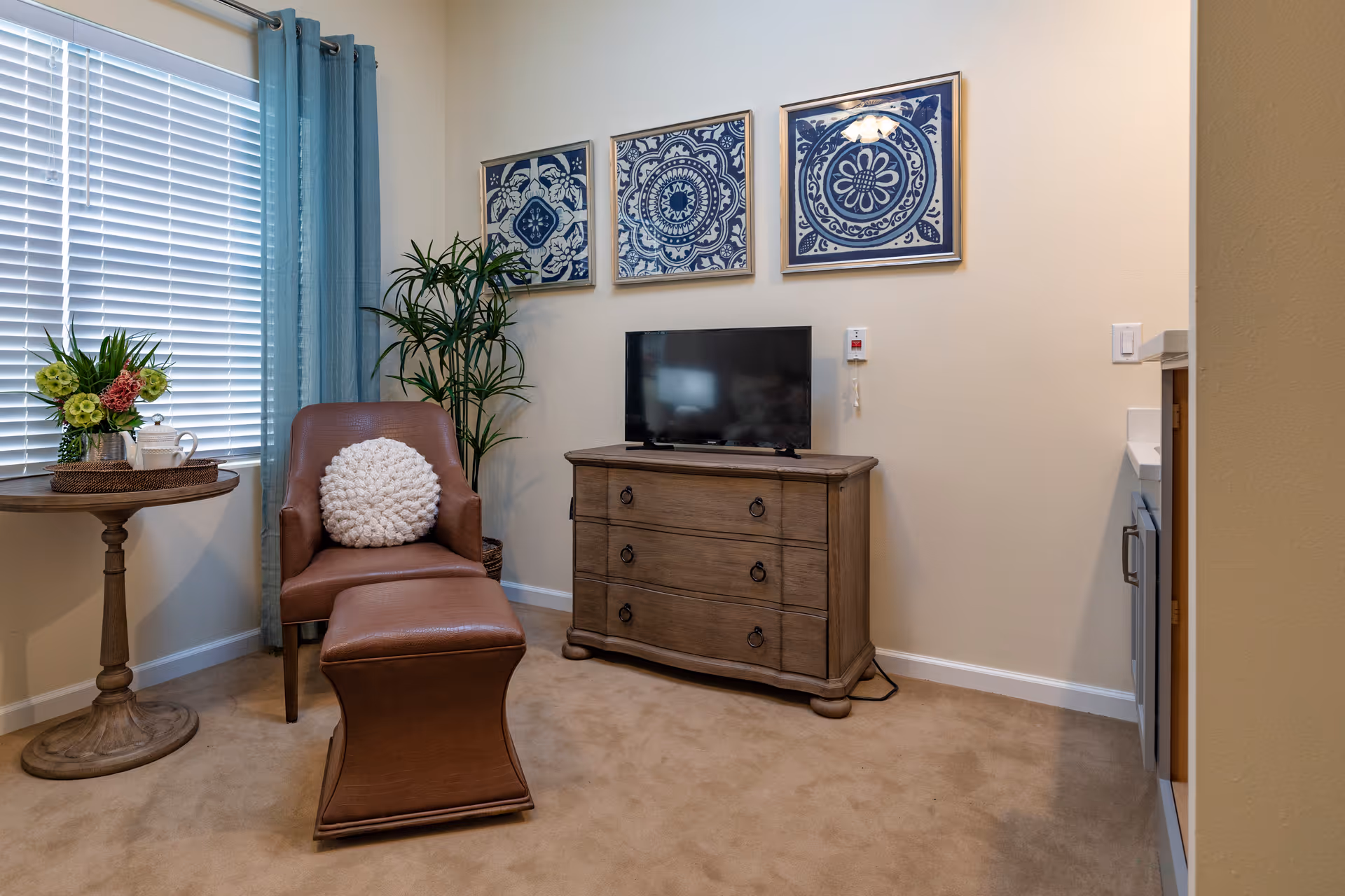 A cozy living room area with a brown leather armchair and matching ottoman, a round wooden side table with a flower arrangement and a teapot, a wooden dresser with a flat-screen TV on top, three framed blue and white decorative artworks on the wall, a tall green plant in the corner, and a window with white blinds and blue curtains.