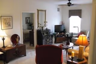 A cozy living room with a red armchair in the foreground, a wooden chest with a lamp and decorative items on the left, a black dresser with a mirror and candles, a TV on a stand, and a beige sofa with a floor lamp next to a window with a valance.