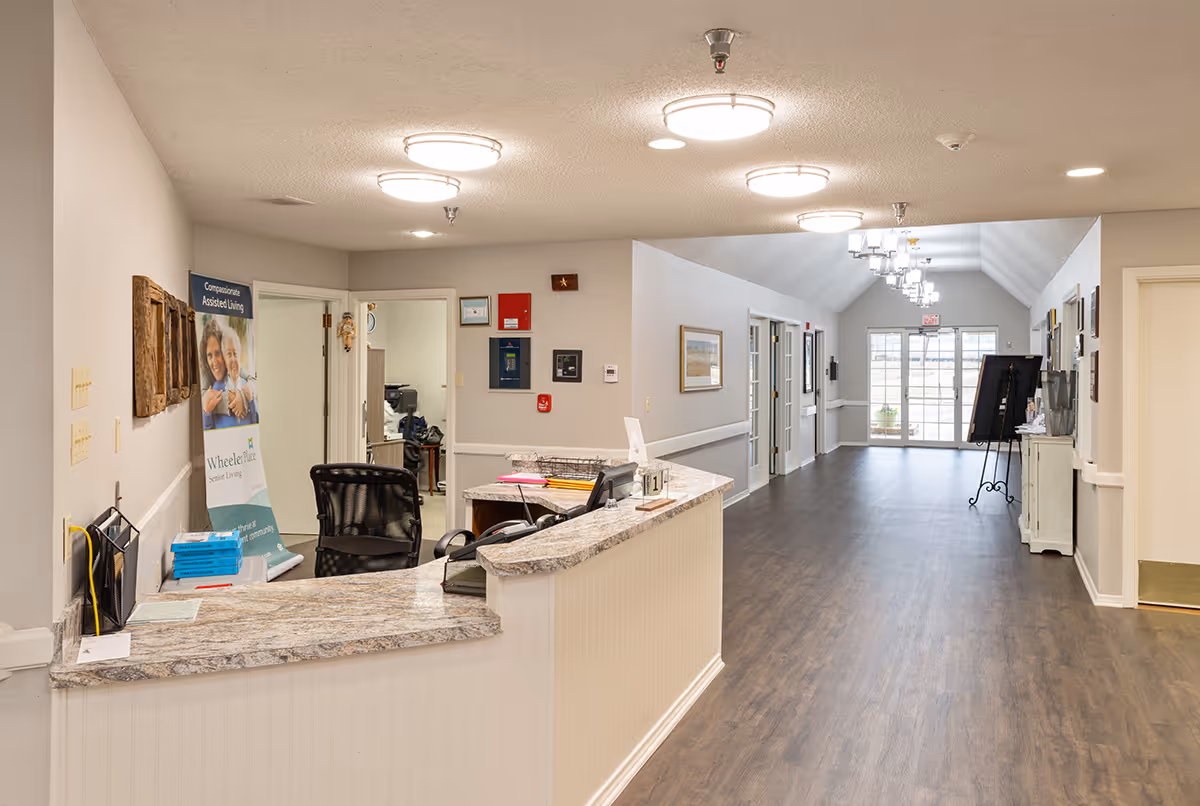 Reception desk and long interior hallway of a senior living facility with a chair, countertop, signage, and doors leading to rooms.
