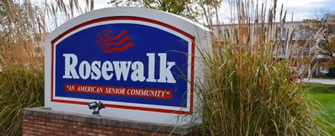 Outdoor view of a large sign for Rosewalk, an American senior community, surrounded by tall ornamental grasses with a building and trees in the background under a partly cloudy sky.