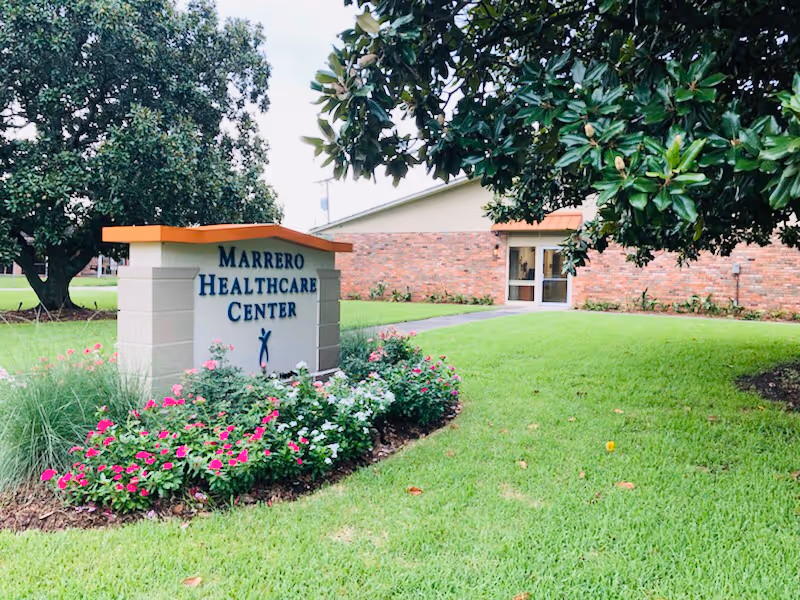 A landscaped lawn and flowerbed with a sign reading "Marrero Healthcare Center" in front of a brick building entrance.