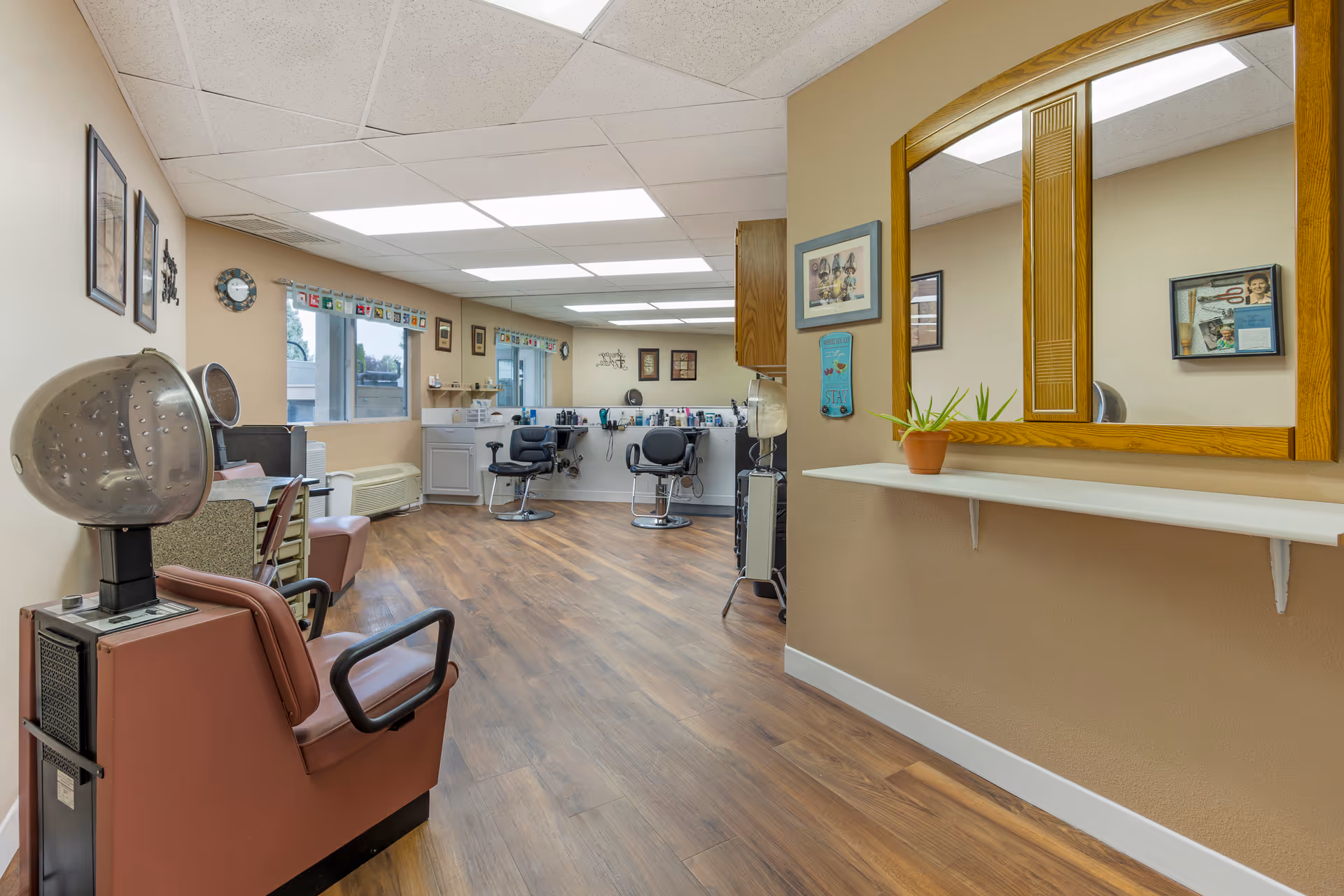 Interior view of a hair salon area in a senior living facility with salon chairs, hair dryers, a large mirror with a wooden frame, and various hair styling tools and decorations on the walls.