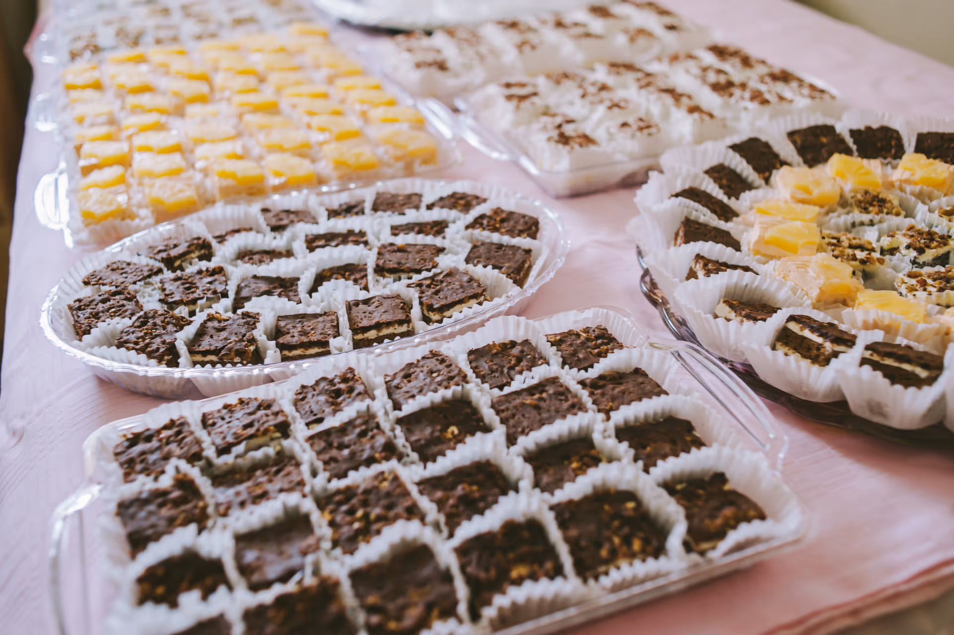 Several trays of assorted small dessert squares, including chocolate and yellow-topped varieties, arranged neatly on a pink tablecloth.