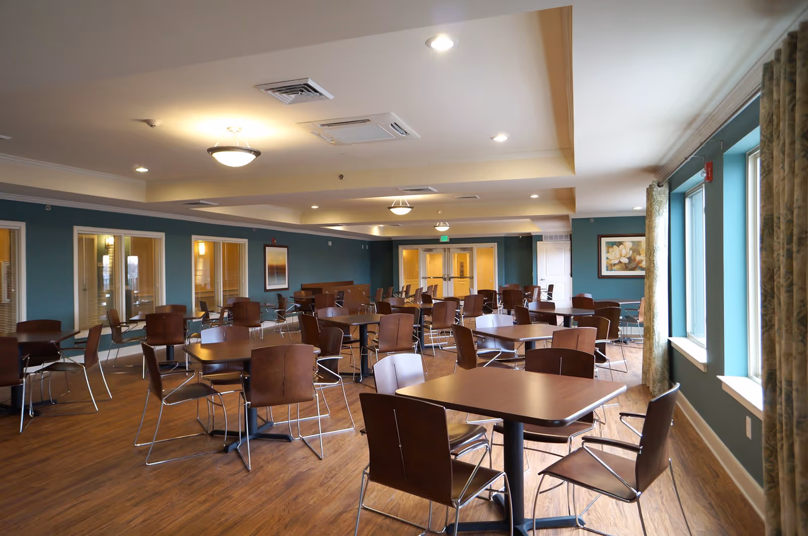 A spacious dining room with multiple square wooden tables and brown chairs arranged neatly. The room has teal walls, large windows with patterned curtains on the right side, wood flooring, and ceiling lights providing warm illumination. There are framed artworks on the walls and glass doors at the far end of the room.