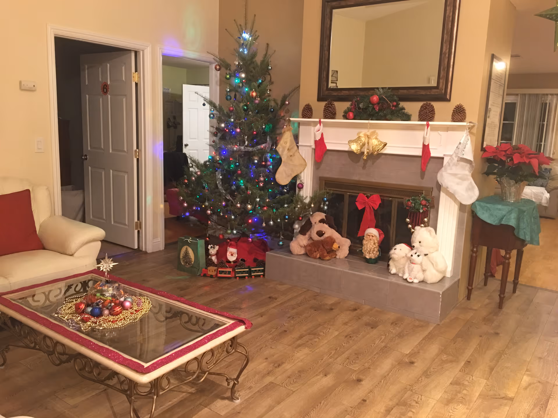A cozy living room decorated for Christmas with a lit Christmas tree adorned with colorful lights and ornaments. There is a fireplace with stockings hanging on the mantel, pine cones, and festive decorations including stuffed animals and a Santa figurine. A glass coffee table with holiday ornaments and a white armchair with a red cushion are also visible. The room has wooden flooring and warm lighting.