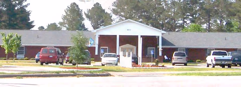 Front exterior of a single-story brick building with a white portico entrance and several parked cars in the driveway.