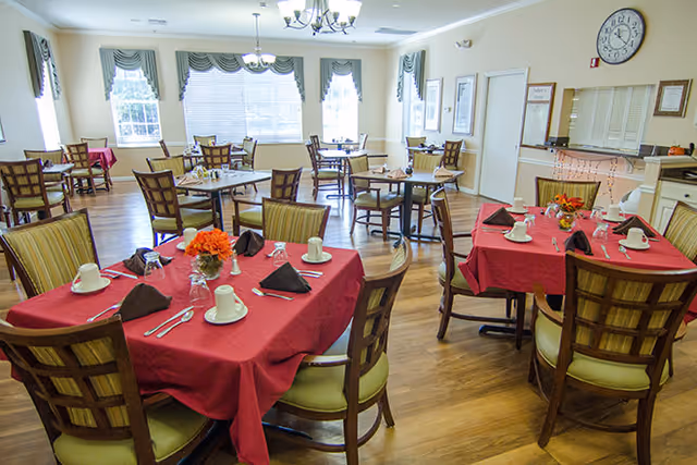 A bright dining room with multiple tables covered in red tablecloths, each set with white cups, silverware, and black napkins. The room has large windows with green curtains, wooden chairs with cushioned seats, and a wooden floor. There is a clock on the wall and framed pictures, creating a welcoming atmosphere.