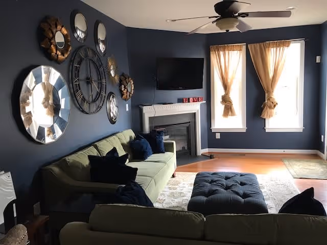 Cozy living room with navy walls featuring decorative mirrors and a large clock, a green sofa, ottoman, fireplace with mounted TV, and two curtained windows.