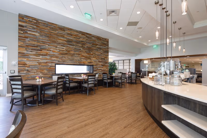 A spacious dining area with wooden flooring and a stone accent wall featuring a horizontal window. Several tables with chairs are arranged along the wall and in the background. On the right side, there is a curved counter with pendant lights hanging above it, and various items placed on the counter. The ceiling has recessed lighting and air vents.