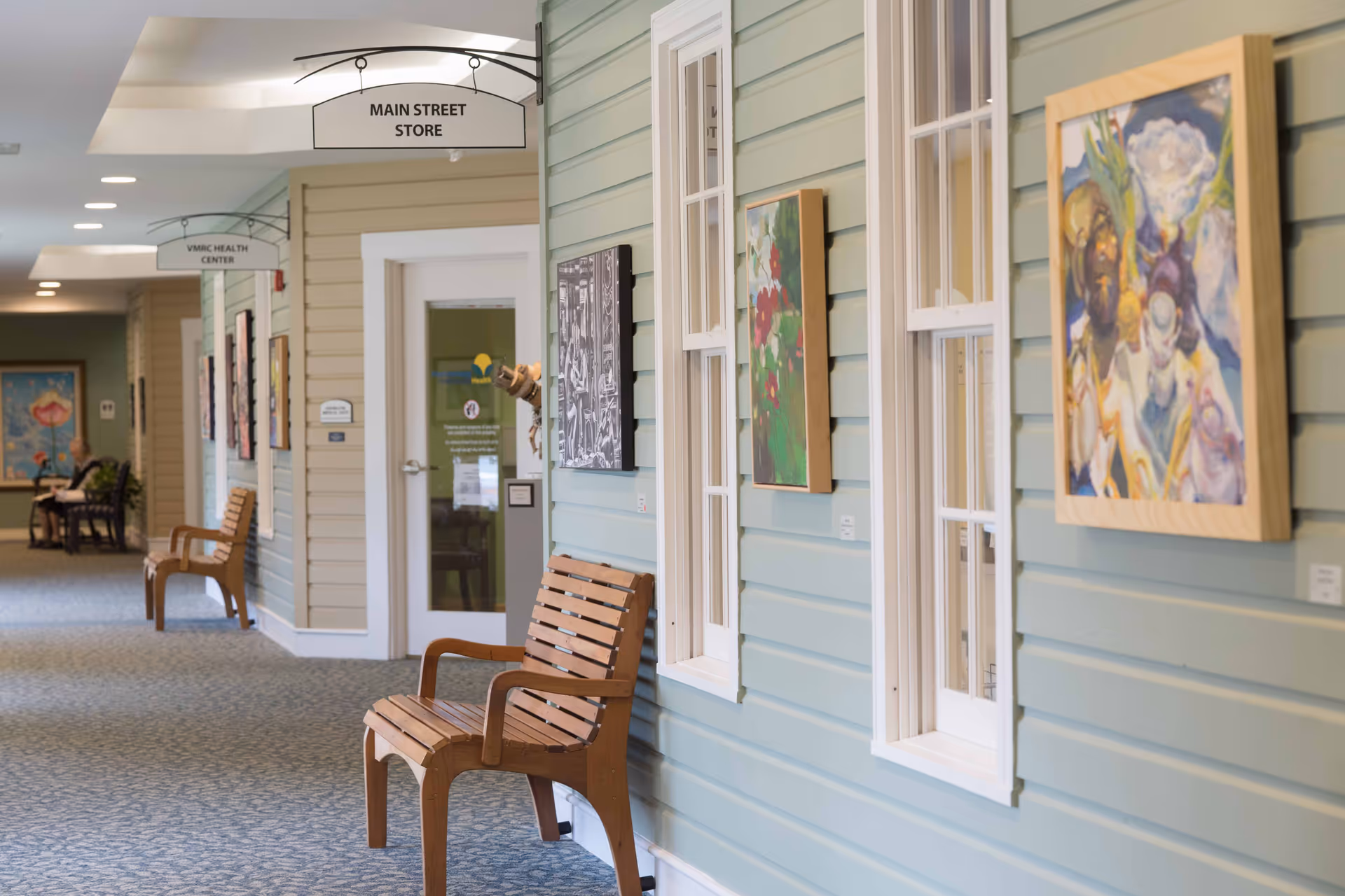 Indoor corridor with wooden bench seating, framed paintings on pale blue siding walls, and overhead directional signs.