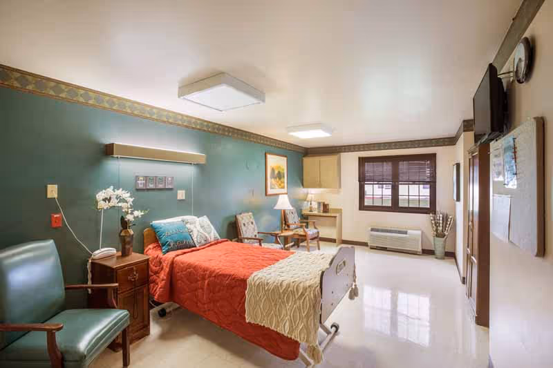 A well-lit senior living bedroom with a single bed covered in a red quilt and a beige throw blanket. The room features a green accent wall with a mounted light above the bed, a wooden nightstand with a vase of white flowers, and two upholstered chairs with a small table and lamp between them. There is a window with brown blinds, a wall-mounted TV, and a heating/cooling unit below the window. The floor is shiny and light-colored, and the room has a calm, cozy atmosphere.