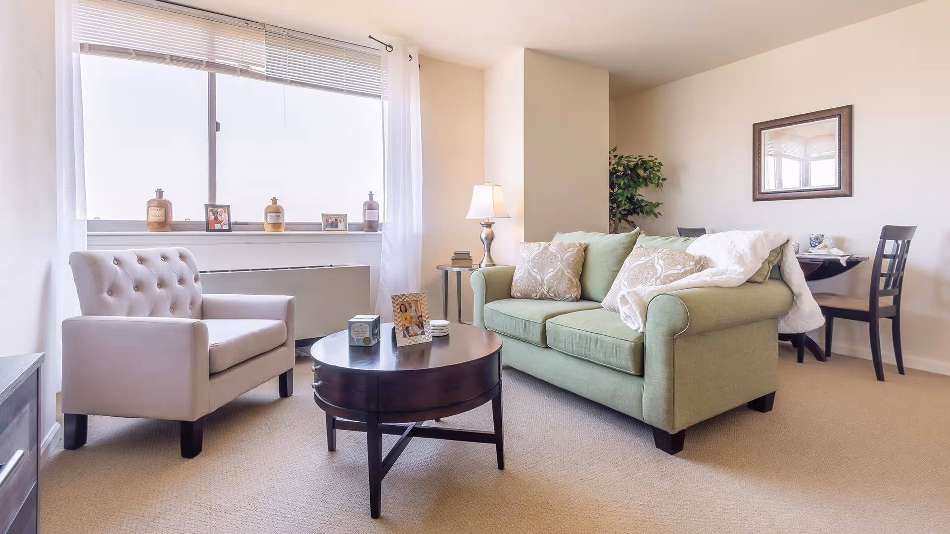 A bright living room with a large window covered by white curtains. The room features a light green sofa with patterned cushions and a white throw blanket, a beige armchair, and a round dark wooden coffee table with decorative items on it. In the background, there is a small dining table with two chairs and a framed mirror on the wall.