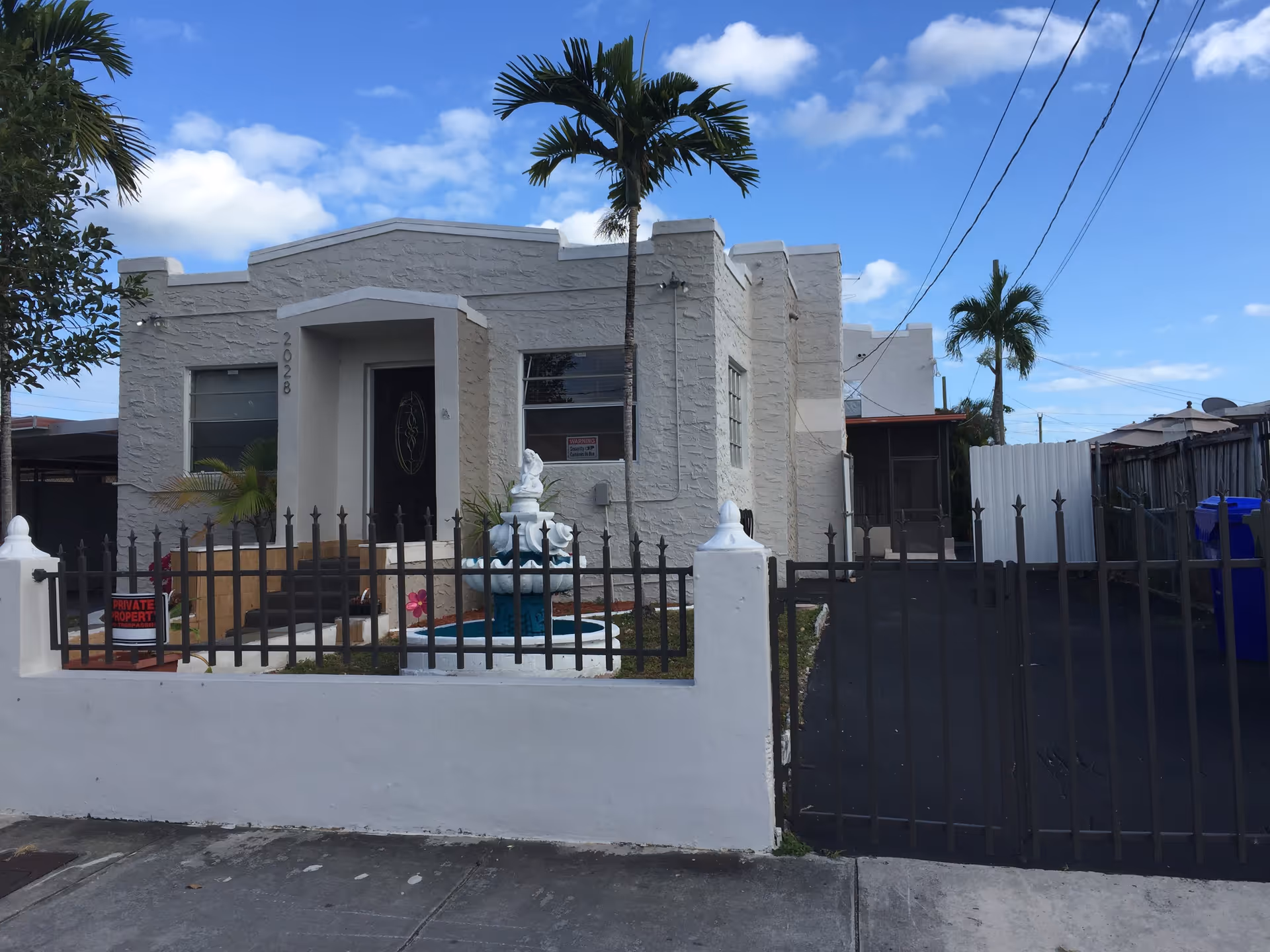 Exterior view of a single-story white stucco building with a gated front yard. The yard features a decorative fountain and some small plants. There are palm trees around the property, and the sky is partly cloudy.