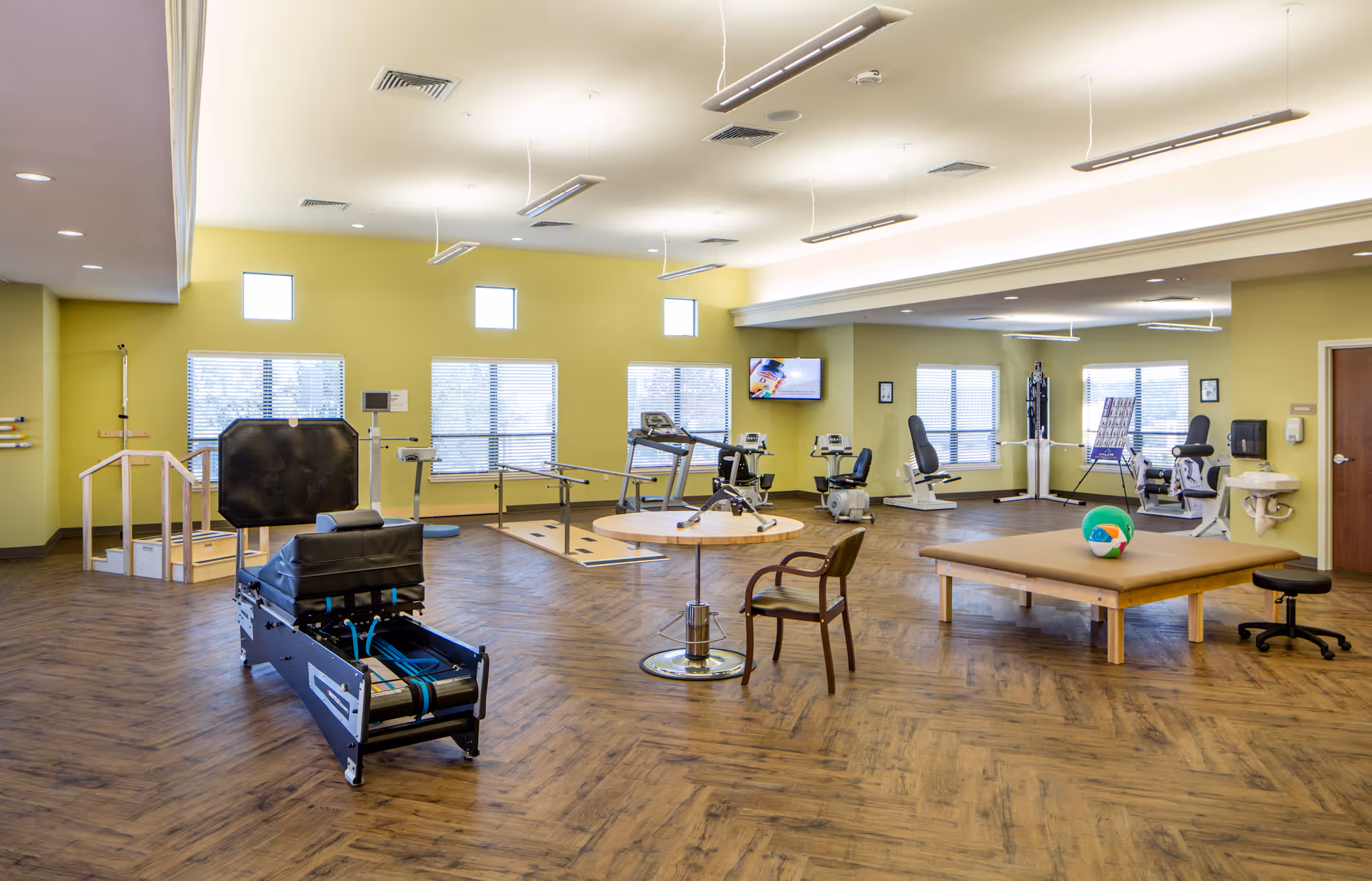 A spacious physical therapy or rehabilitation room with exercise equipment including parallel bars, stationary bikes, a therapy table with a colorful ball, a chair, and a specialized exercise machine. The room has large windows with blinds, light green walls, wood-patterned flooring, and ceiling lights.