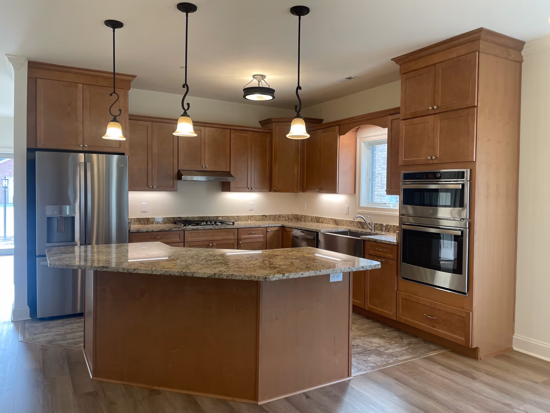 Modern kitchen with wooden cabinets, granite countertops, stainless steel refrigerator, double oven, dishwasher, and a large island with three pendant lights hanging above it.