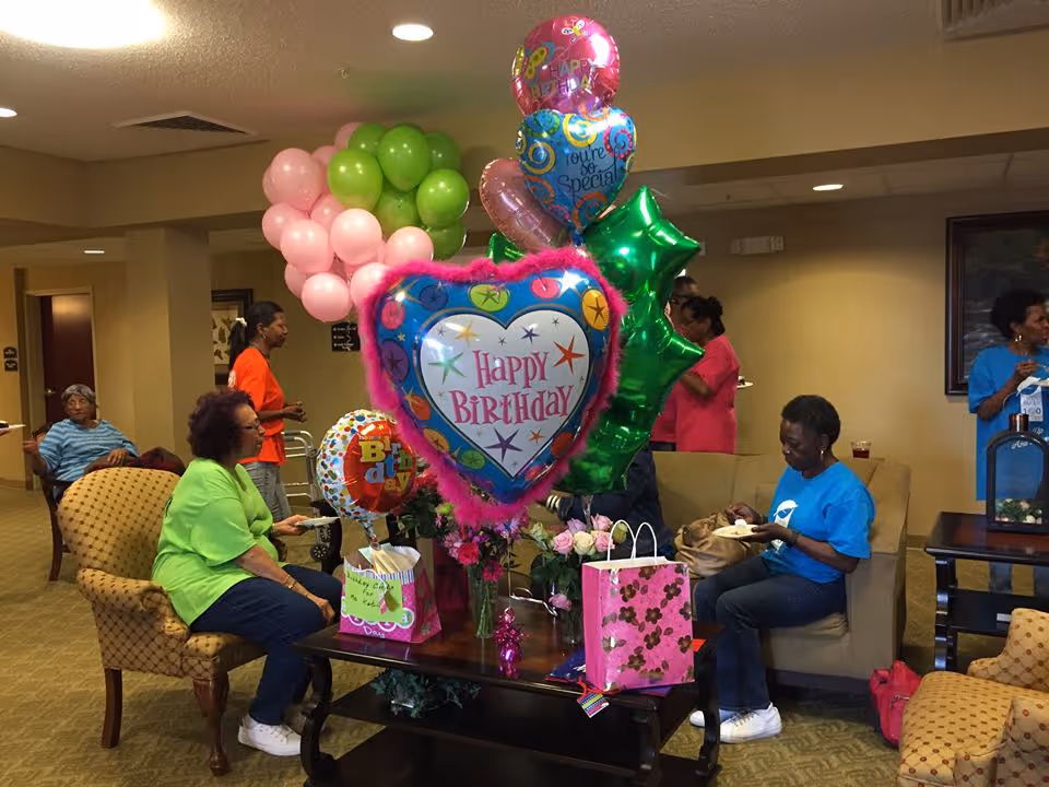 Residents gather in a senior living common room decorated with 'Happy Birthday' balloons and gift bags.