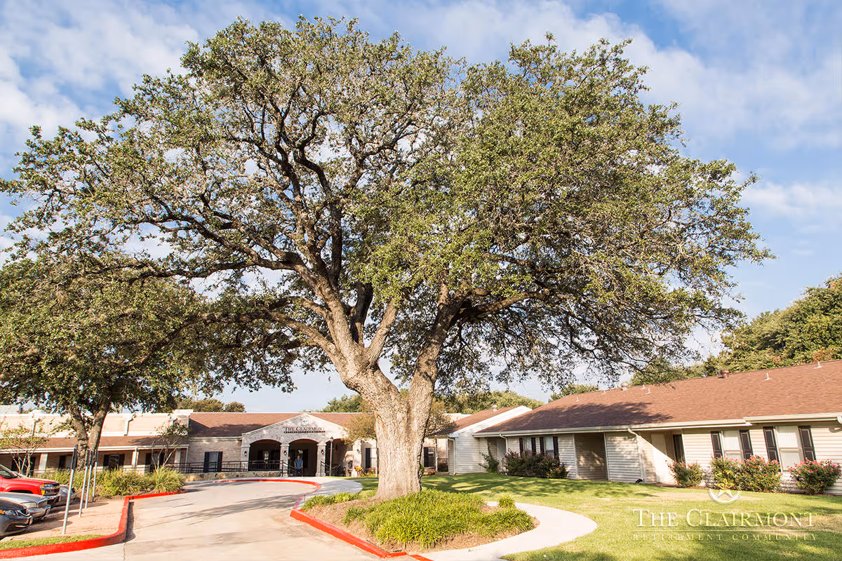Exterior view of The Clairmont Retirement Community building with a large tree in the foreground, a curved driveway, and parked cars on the left side under a partly cloudy sky.