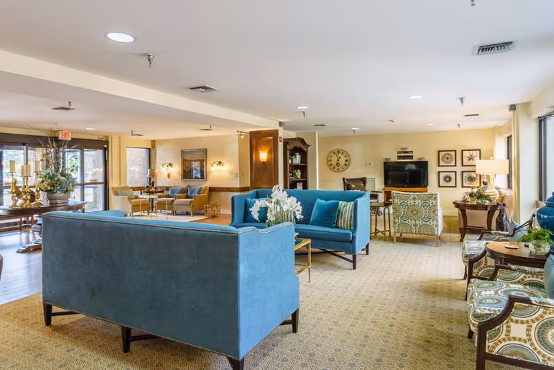A spacious and well-lit living room area in University Place Independent Living featuring blue upholstered sofas, patterned armchairs, a television on a wooden stand, decorative wall art, and various tables with lamps and floral arrangements. Large windows allow natural light to fill the room.