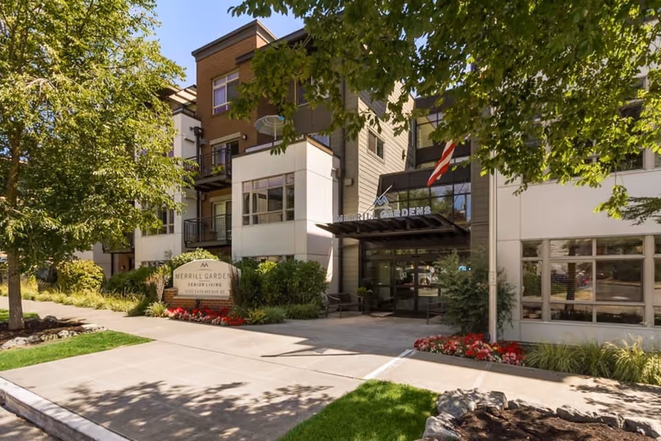 Entrance and landscaped front of the Merrill Gardens at The University senior living building with a sign, walkway, and American flag.