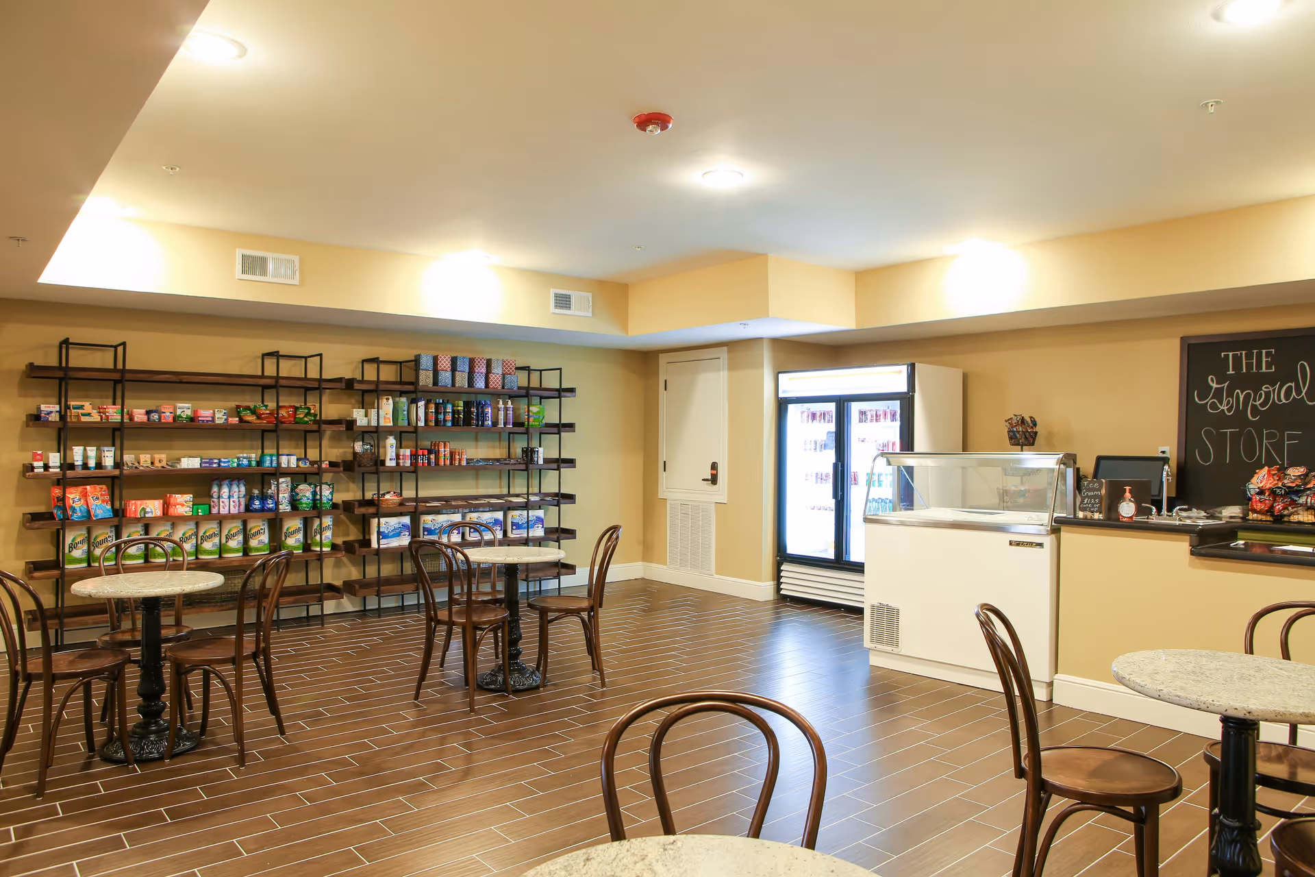 Interior view of a small store area in a senior living facility with shelves stocked with various snacks and household items, a refrigerated display case, small round tables with chairs, and a chalkboard sign that reads 'The General Store'.