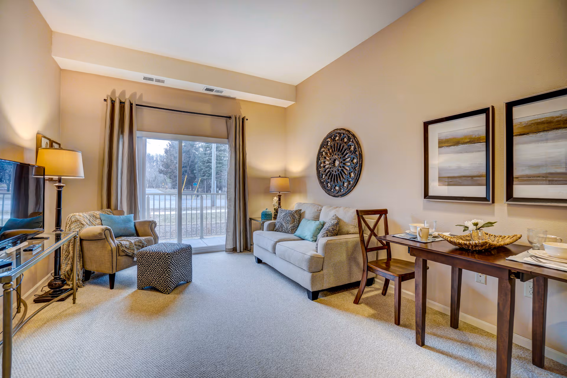 A cozy living room with a beige sofa adorned with decorative pillows, a patterned ottoman, and an armchair with a blue cushion. There is a glass TV stand with a flat-screen TV on the left, two framed abstract paintings on the right wall, and a small dining table set for two. A large sliding glass door with curtains lets in natural light and offers a view of trees outside.