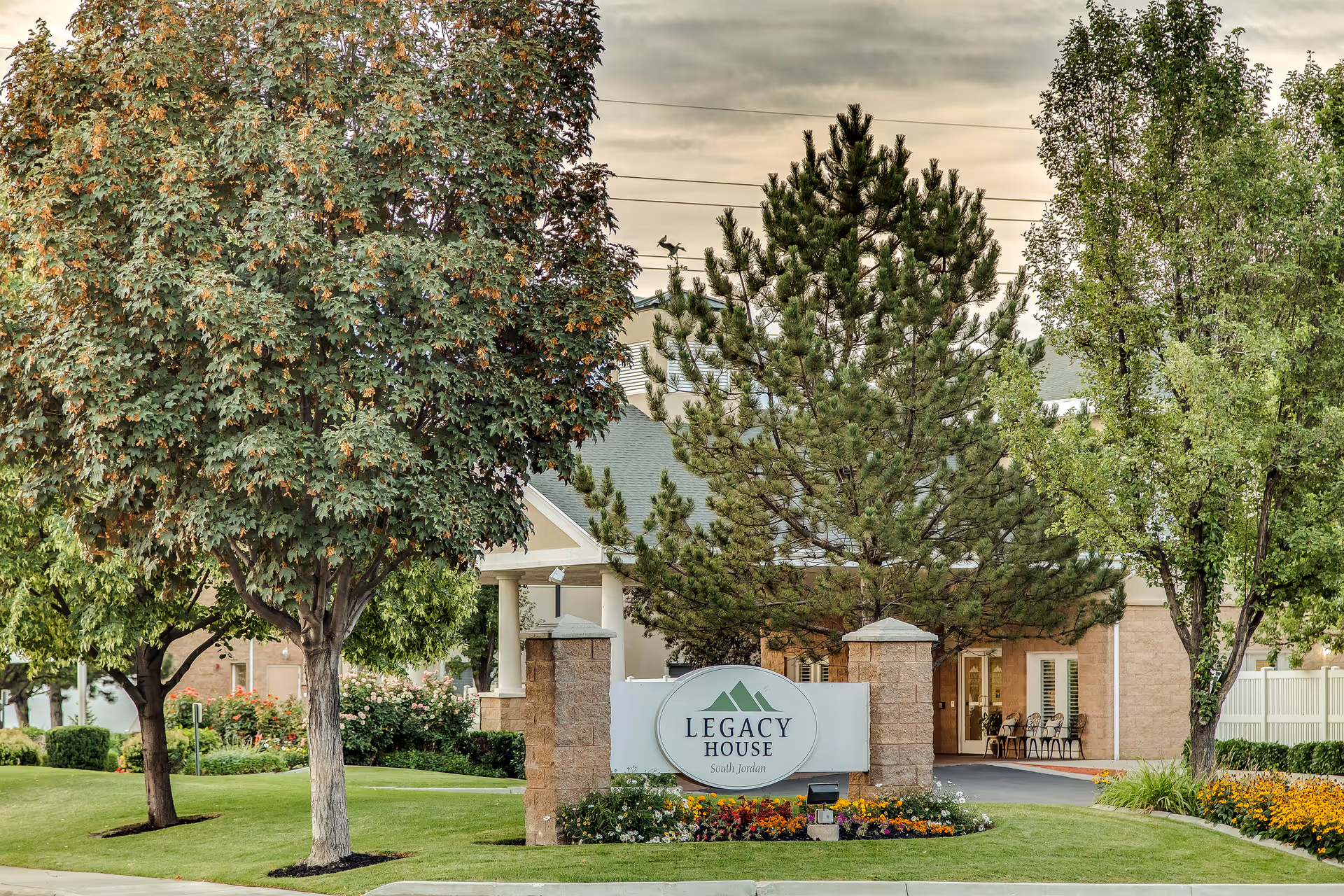 Exterior view of Legacy House of South Jordan facility with a landscaped lawn, trees, and a sign displaying the facility name in front of the building.