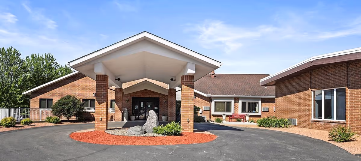 Front entrance of a single-story brick senior living building with a covered porte-cochère, circular driveway, and landscaping.