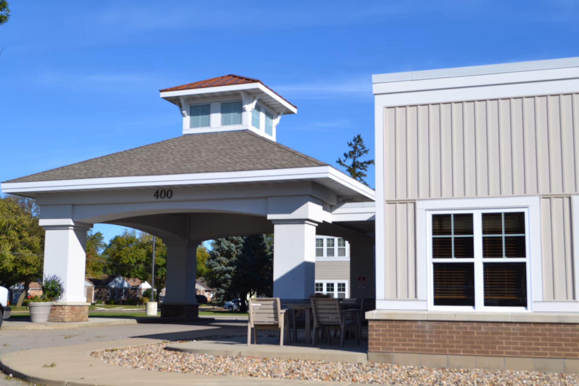 Exterior view of a building entrance with a covered driveway supported by large white columns. The building has beige siding with a brick base and a window with white trim. The number 400 is displayed above the entrance. There are outdoor chairs and a table near the entrance, and trees and houses are visible in the background under a clear blue sky.