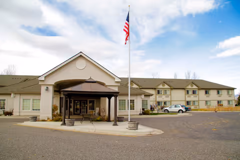 Exterior view of a senior living facility building with a covered entrance, an American flag on a flagpole, and a parking area with a few cars. The sky is partly cloudy.