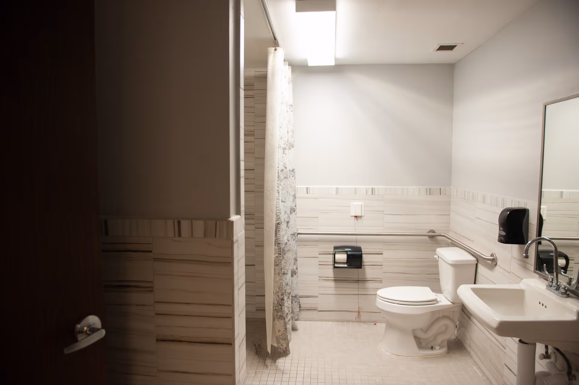 A clean, accessible bathroom with a toilet, sink, and a shower area with a curtain. The walls are tiled halfway up with light-colored tiles, and there are grab bars installed near the toilet for support. A mirror and a soap dispenser are mounted above the sink.