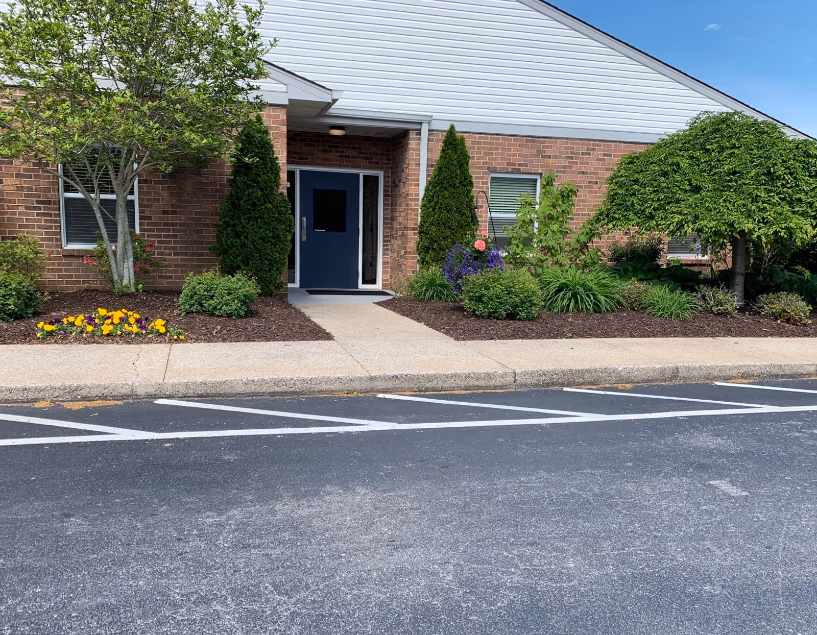 Front entrance of a brick building with a blue door, landscaped flower beds and a parking area.