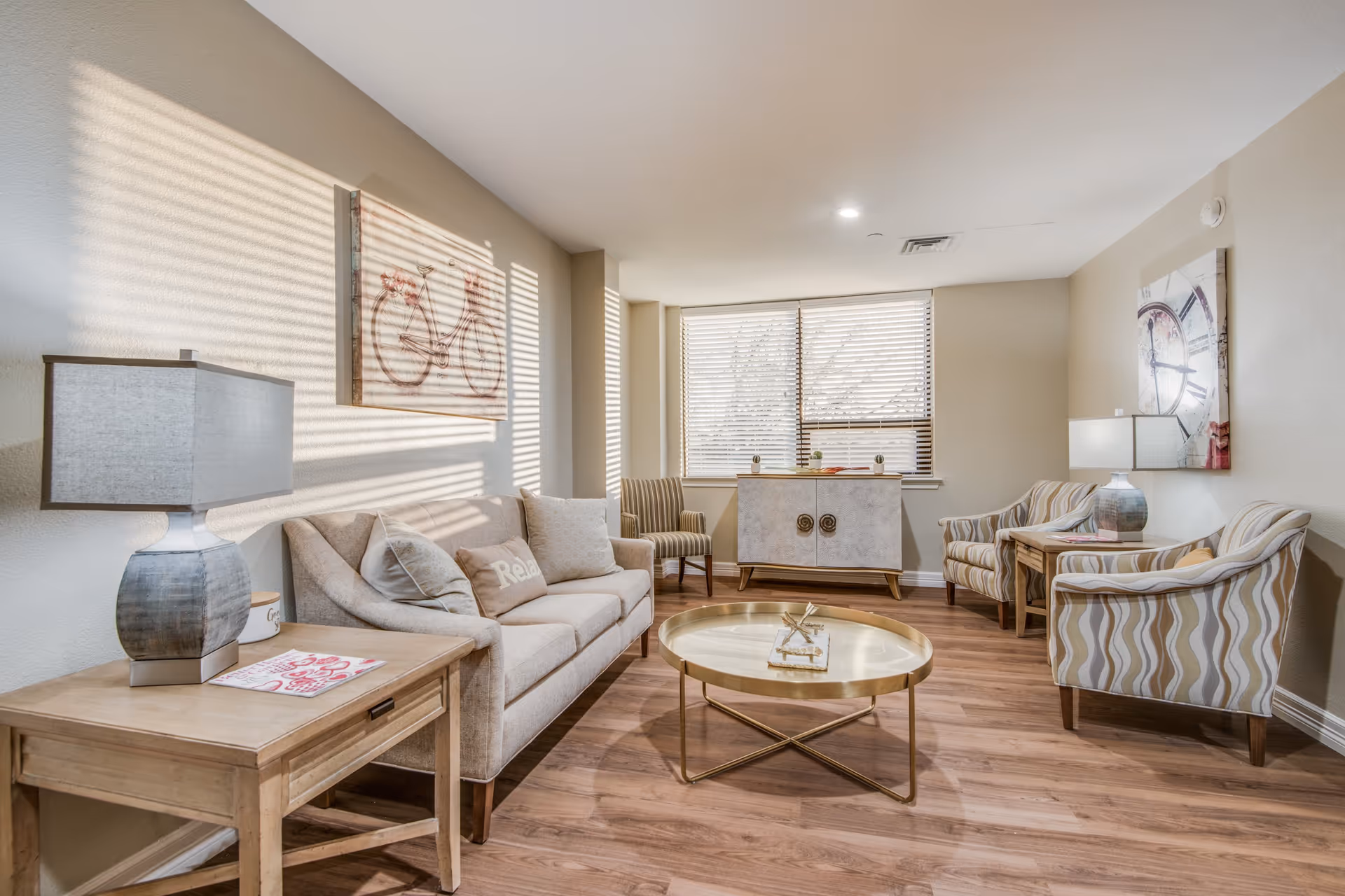 A bright and cozy living room with a beige sofa adorned with pillows, two striped armchairs, a round gold coffee table, and two wooden side tables with lamps. The room has wooden flooring, a large window with blinds, and wall art including a bicycle and a clock.