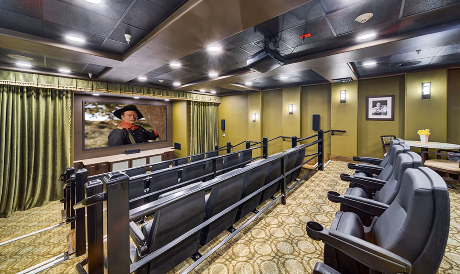 A small theater room with rows of black cushioned seats facing a large screen displaying a man in a hat. The room has green curtains on the sides of the screen, patterned carpet flooring, and green walls with framed pictures and wall sconces providing lighting.