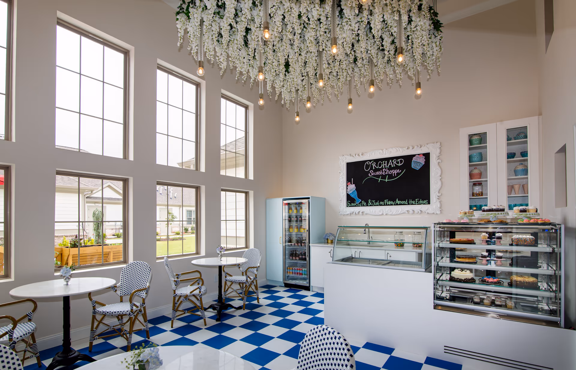 Bright and airy sweet shop area with large windows, blue and white checkered floor, white round tables with patterned chairs, a display case filled with cakes and cupcakes, a refrigerator stocked with drinks, and a decorative ceiling with hanging white flowers and light bulbs. A chalkboard sign reads 'Orchard Sweet Shoppe'.
