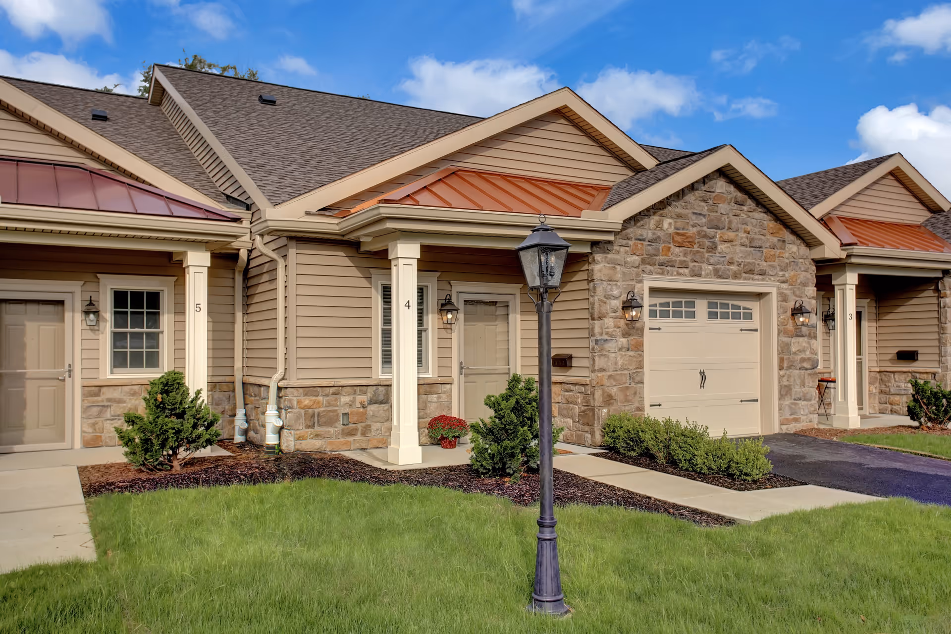 Front exterior of connected single-story residential units with stone accents, garages, a lamppost, and a manicured lawn.