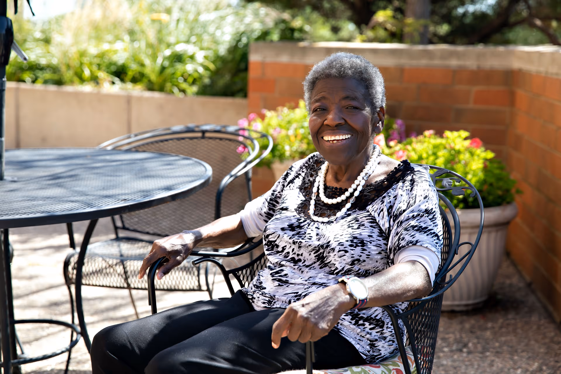 An elderly woman sitting outdoors on a metal chair at a patio table, smiling warmly. She is wearing a patterned black and white top, black pants, a pearl necklace, and a watch. Behind her are potted plants and a brick wall.